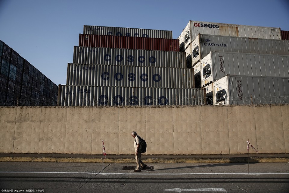 A pedestrian passes stacked COSCO Pacific Ltd. branded shipping containers behind a perimeter wall in the commercial shipping area of Piraeus Port, operated by Piraeus Port Authority SA, in Piraeus, Greece, on Monday, May 18, 2015. A sale of the port would be a reversal on the part of Greece's Syriza party-led government, which had earlier pledged to block such moves. Photographer: Yorgos Karahalis/Bloomberg
