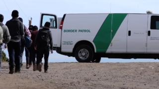 A Border Patrol van parked with the rear doors open. And a group of migrants lining up to get in.