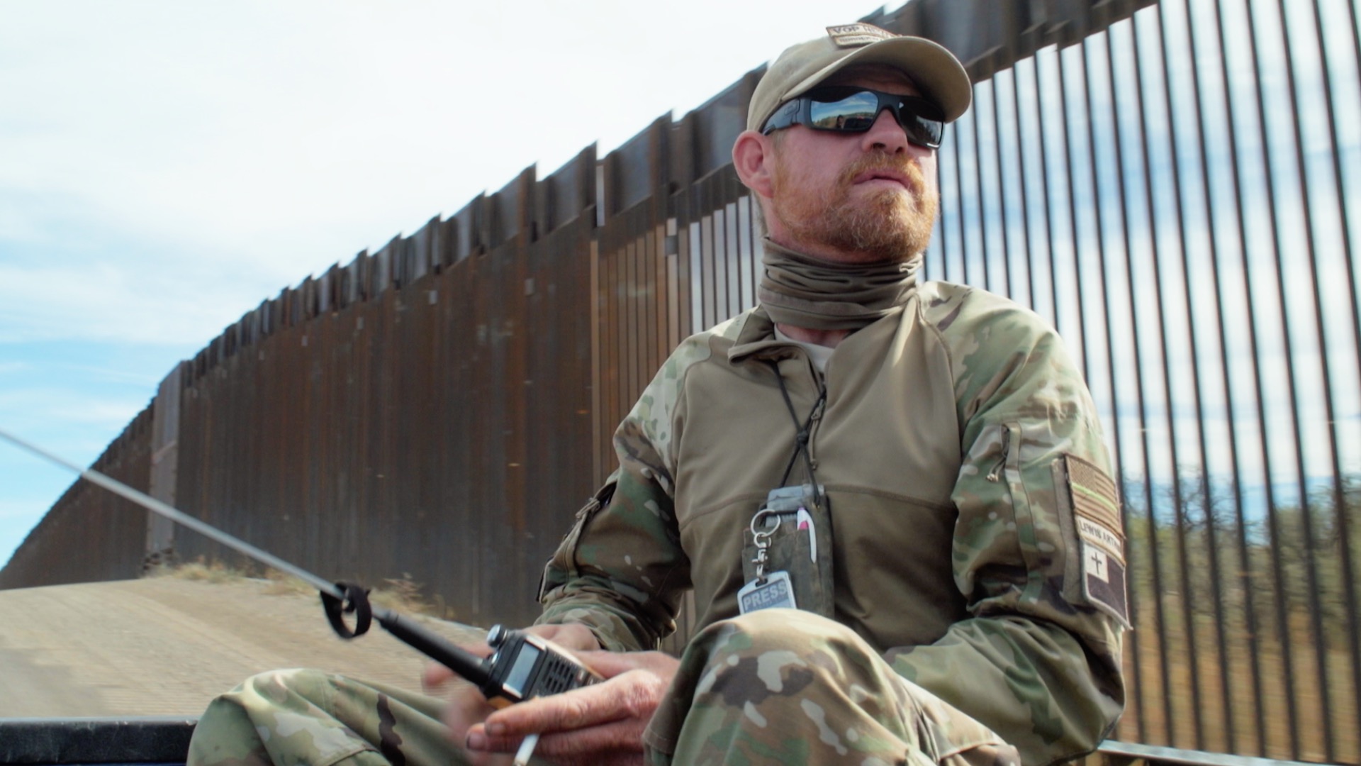 An armed vigilante sits on the back of a truck patrolling the U.S–Mexico border. You can see the wall dividing the countries behind him.