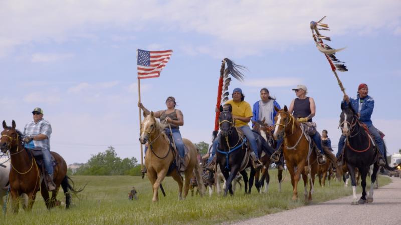 A group of Lakota horse riders, some carrying flags and in Lakota regalia.