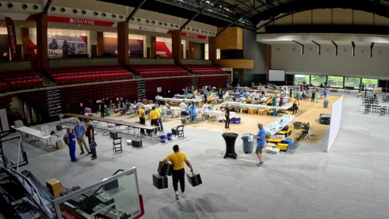 A group of people are setting up a mobile clinic inside the gymnasium of the University of Virginia at Wise. There are gurneys, assorted equipment, and volunteers wearing yellowy T-shirts carrying boxes.