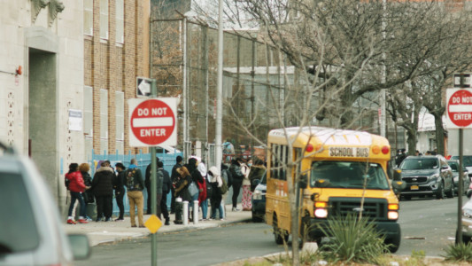 A school bus leaves the stop as a group of students is seen on the sidewalk in front of the school building.