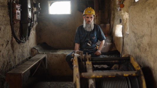 A miner with a long white beard, a yellow helmet and a denim overall sits in front of a piece of mining machinery.