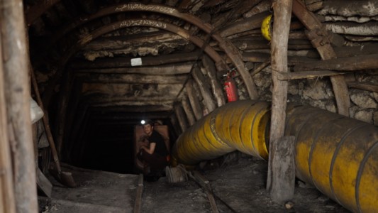 A mining tunnel with wooden beams and a large yellow duct alongside its lower right side. At the end of the shot, a miner is staring back. He is barely visible, mainly because to the light on his hat.