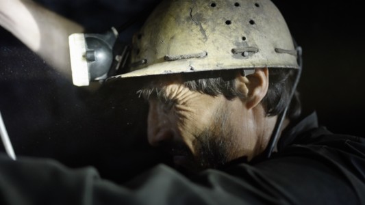 The head of a miner seen from the side. His face is covered in mud, as is his helmet.