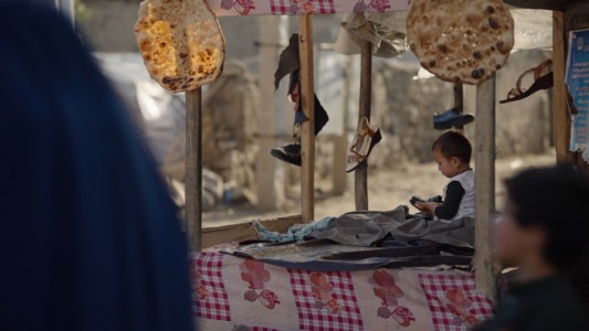 A baby boy sits on a street stand, shoes are hanging from its poles. We also see loaves of bread in display from the contiguous stand.