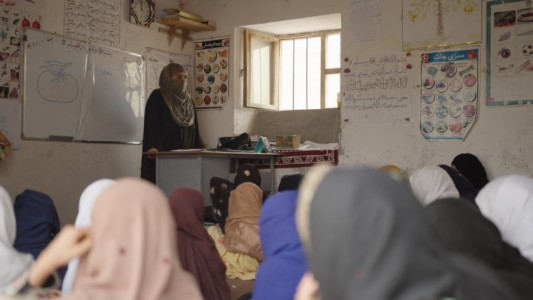 Inside a classroom in Afghanistan, a teacher stands in front of her students. We only see the head covers of the students, as the teacher is facing the camera.