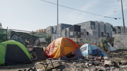 Three tents on the forefront, surrounded by debris and trash, a fence separating them from a modern development in the back.