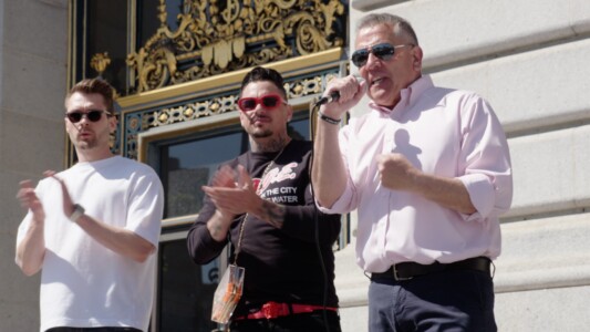 Three people in front of San Francisco City Hall during a protest. One of them is holding a microphone. The other two are clapping.