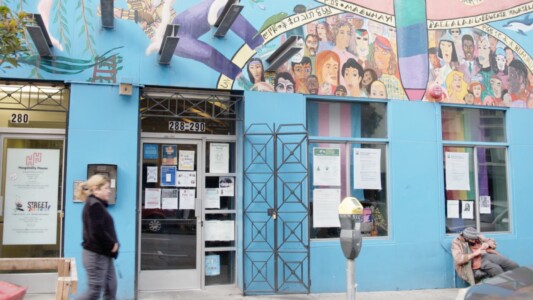 The front of a building. It is painted light blue and has a mural on top of its door and window. A person is walking by and someone is sitting in a chair.